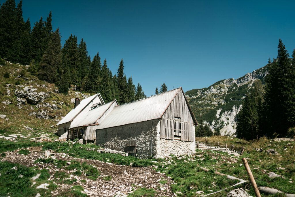 A traditional stone and wooden alpine barn in a grassy meadow with the rocky peaks of the Julian Alps in the background near Lake Krn.

